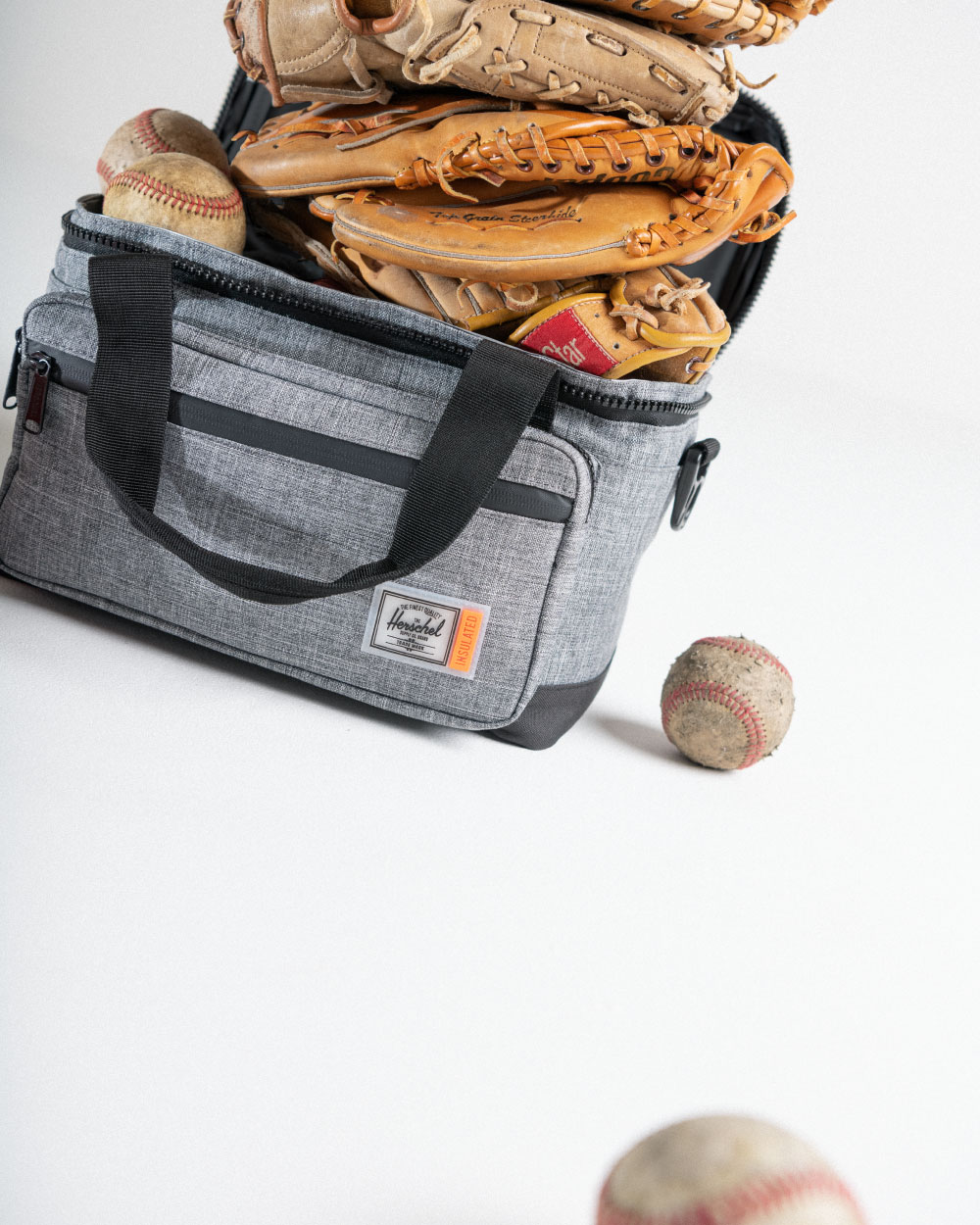 Three baseball gloves and some baseballs sticking out of the main compartment of the Herschel Supply Pop Quiz Cooler 12 Pack Insulated in Raven Crosshatch